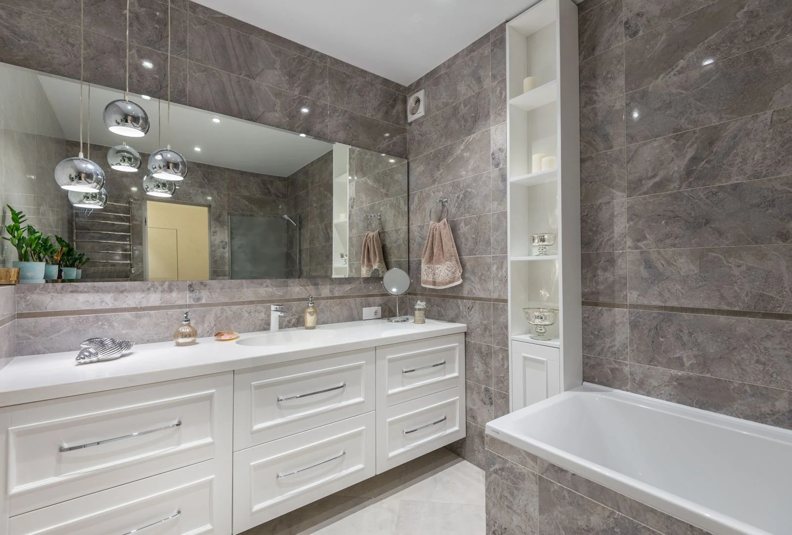 Big window hanging on tiled wall above while cabinet and sink near bathtub in modern bathroom decorated with creative lamps and potted plants