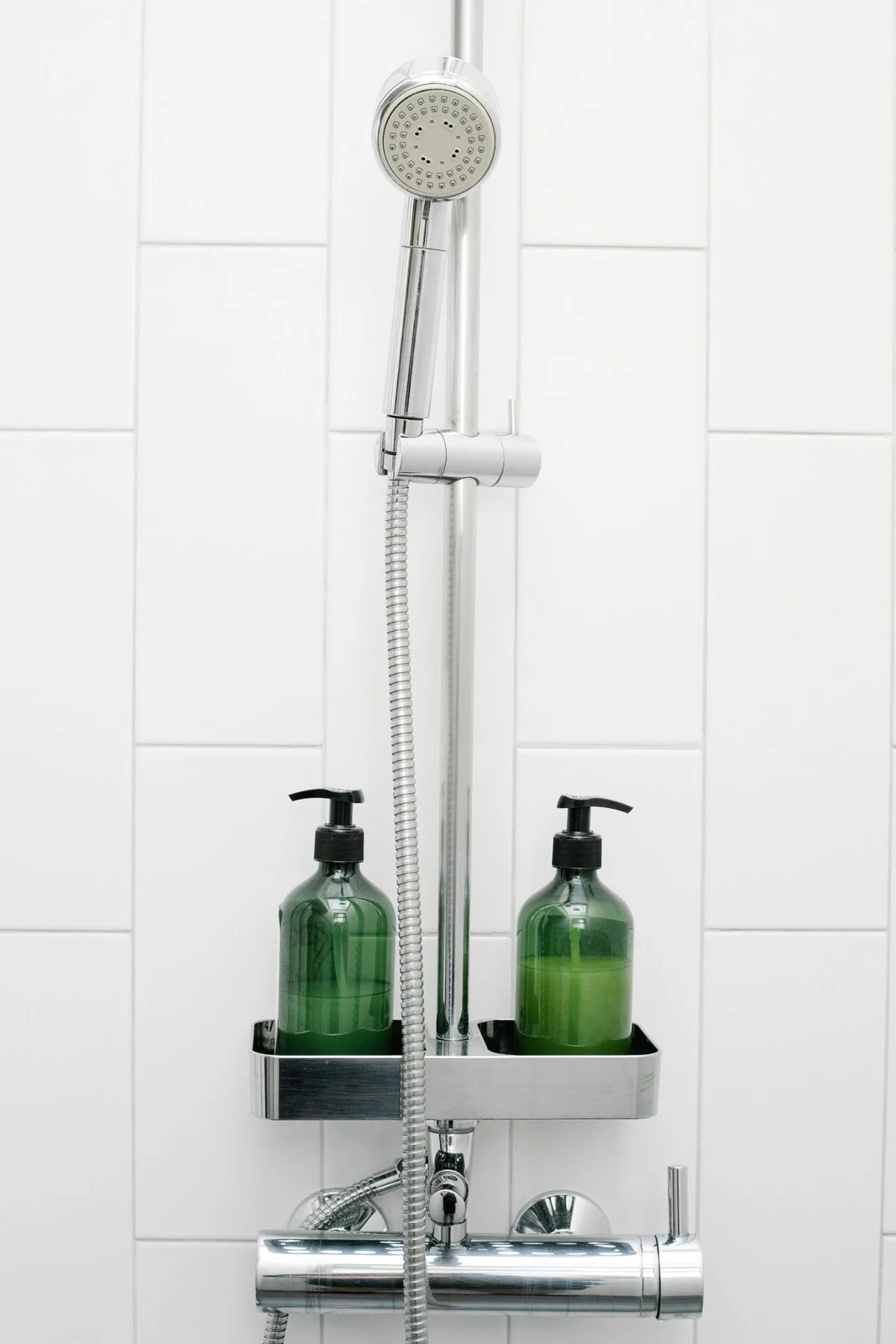 Handshower and hose attached to white tiled wall near bottles with essential skin care products in light bathroom